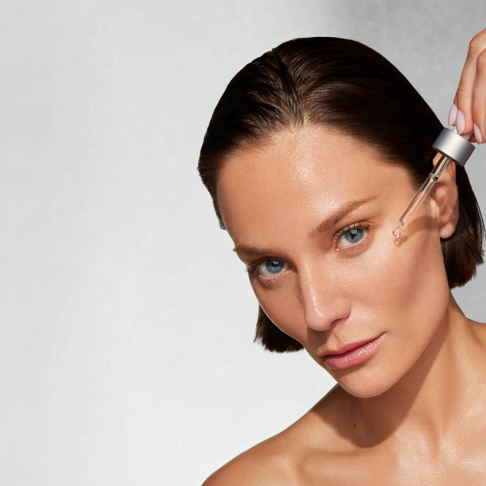 Frau mit erhobenem Arm trägt per Pipette Serum aufs Gesicht.